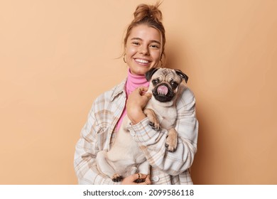 Happy Female Pet Owner Dressed Casually Holds Pug Dog Which Sticks Tongue Spends Free Time With Favorite Puppy Isolated Over Beige Studio Background People Animals And Relationship Concept