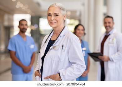 Portrait Of Mature Female Doctor On Hospital Corridor Confident General Practitioner Standing In Hospital Hallway With Her Healthcare Team In Background Successful Head Physician In Private Clinic